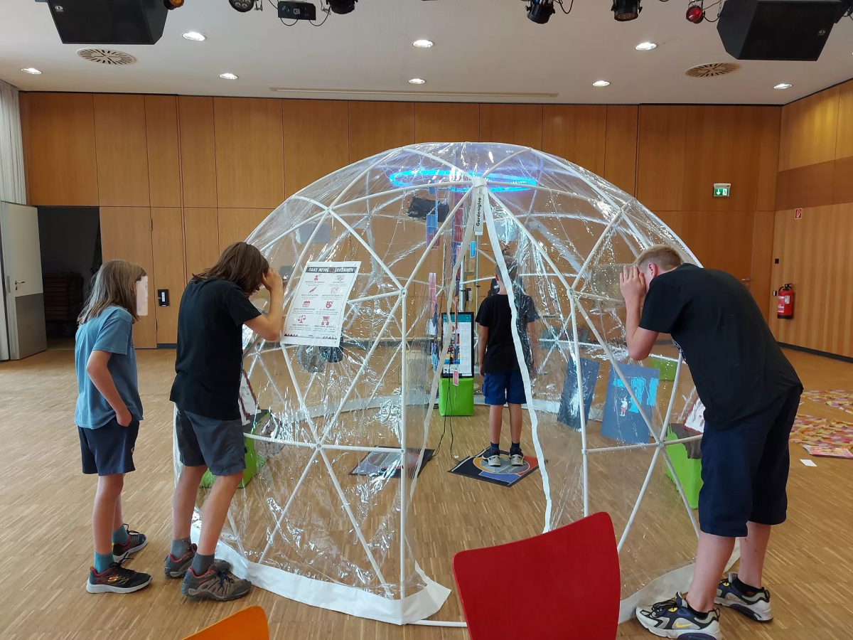 Jugendliche erkunden von innen und von außen das transparente Kuppelzelt in der Ausstellung „Cyberbubble“ und informieren sich zum Thema Fake News.